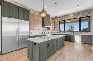 A modern kitchen with green cabinetry and a central island featuring a white countertop. The kitchen is equipped with a large stainless steel refrigerator, built-in oven, and dishwasher. Pendant lights hang above the island, and a large window offers an outdoor view.