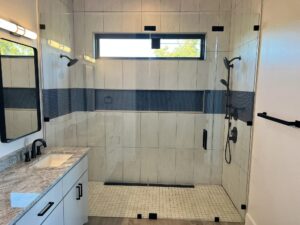 A modern bathroom features a walk-in shower with a horizontal window, light-colored tiles, and black fixtures. The vanity has a granite countertop, white cabinets, and a rectangular mirror above. A black towel rack is mounted on the wall.