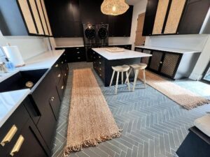 Stylish modern laundry room with dark cabinets, gold hardware, two black front-loading washing machines, natural woven rugs, white countertops, a central island with two stools, and a pendant light made of woven material. The floor has a herringbone pattern.