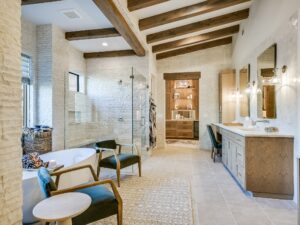 A luxurious bathroom with exposed wooden beam ceiling features a free-standing tub, glass-enclosed shower, double vanity with twin mirrors and modern lighting. Two green armchairs and a small round table sit on a textured rug near the tub. A walk-in closet is visible.