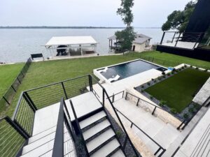 View from a modern balcony overlooking a backyard with a pool, green lawn, and a lakeside boat dock with two covered structures. The serene lake stretches across the background, encapsulating a peaceful, waterside ambiance.