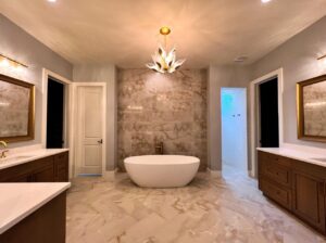 A luxurious bathroom featuring a central freestanding white bathtub against a marble wall. Two wooden vanities with white countertops and framed mirrors are on either side. A gold and glass chandelier hangs from the ceiling, illuminating the space.