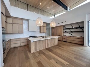 A modern kitchen with wooden cabinets, white countertops, a central island with a sink, and wooden flooring. Two large woven pendant lights hang above the island. The back wall features tile backsplash, an oven, and open shelves on the right side.