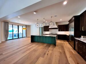 A modern kitchen and dining area with wooden flooring and an island with a teal base and a white countertop. The kitchen features dark cabinets, a stainless steel refrigerator, three pendant lights, and large sliding glass doors leading to an outdoor area.