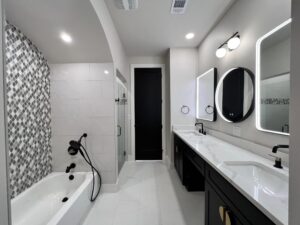 A modern bathroom featuring a bathtub with black fixtures and geometric tile backsplash, a shower with glass door, double sink vanity with illuminated mirrors, black cabinetry, and sleek lighting fixtures. The color scheme is predominantly white, gray, and black.