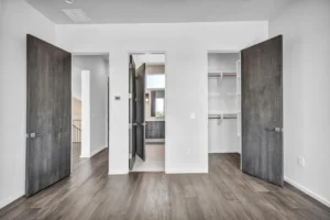 A bright, modern room features light wood flooring and white walls. The room has four dark wooden doors; one open door reveals a bathroom with a vanity, another reveals a closet with built-in shelves. The space shows an entrance leading to a hallway and staircase.