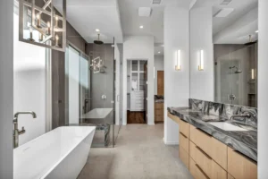 Modern bathroom with a freestanding bathtub, glass-enclosed shower, and gray walls. Double sinks with a marbled countertop are on the right, under two wall-mounted lights. Hanging light fixtures add to the contemporary feel. A walk-in closet is visible at the back.