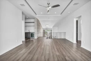 A spacious, modern open-concept living area with light gray walls and dark wood flooring. The ceiling features recessed lighting and a sleek, stainless steel ceiling fan. The area in the back has a small bar with a sink and wine cooler, and a staircase with metal railing leads to the lower floor.