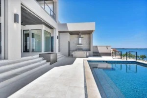 A modern, white concrete house overlooking a large body of water features an outdoor patio with a barbecue area. Stairs lead from the patio to a sparkling rectangular swimming pool under a clear blue sky. Large glass doors connect the interior to the patio.