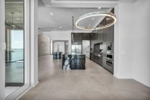 A modern, minimalist kitchen with a large black marble island, dark cabinets, and stainless steel appliances. A unique circular light fixture hangs above the island. Large windows offer a view of the water, and the space is open and airy with light-colored flooring.