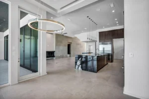 A modern, minimalist kitchen and living space with large windows, light gray floor tiles, a dark marble island, hanging lights, and recessed ceiling lighting. The area is spacious with a built-in fireplace and an open-concept design.