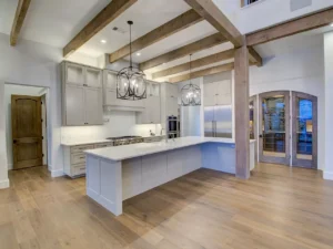 A spacious modern kitchen with light wood flooring, gray cabinetry, and white countertops. Exposed wooden beams cross the ceiling. Three pendant lights hang over the large island, featuring a built-in stove and an oven. A stainless steel refrigerator is in the corner.