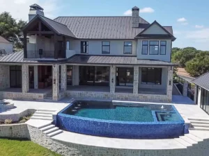 A large, modern two-story house with a combination of stone and wood exterior. A covered balcony and patio are visible. The backyard features a rectangular infinity pool with a blue tile design, surrounded by a spacious stone-paved patio and steps leading down to a lawn.