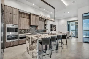 A modern kitchen with high-end appliances, wood cabinetry, and a large marble island with seating for four. A contemporary dining area with a chandelier is visible to the right, and large windows provide natural light, offering a view of an outdoor patio area.