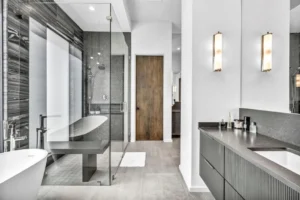 A modern bathroom featuring a freestanding white bathtub beside a glass-enclosed shower. The shower has a dark tile backsplash. The vanity area includes a large mirror, two wall-mounted light fixtures, and a countertop with various toiletries.