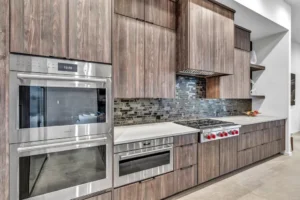 A modern kitchen features stainless steel appliances, including a double oven, microwave, and a gas cooktop with red knobs. The cabinetry has a wood grain finish, and the backsplash is made of dark, reflective tiles. The countertops are white with a smooth finish.